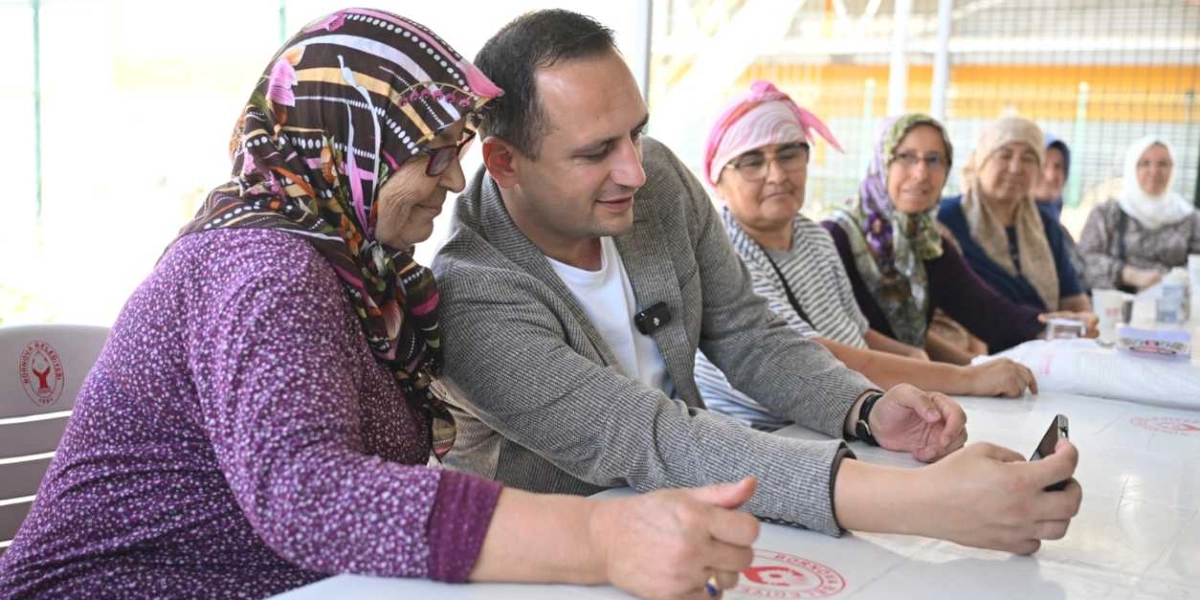 Gri ceketli bir erkek, Bornova Belediyesi logolu beyaz bir masanın etrafında oturan başörtülü yaşlı kadınlara bir akıllı telefon üzerinden bir şeyler gösteriyor. Kadınlar ilgiyle ekrana bakarken, yanındaki yaşlı kadın telefonu tutmasına yardımcı oluyor. Arka planda benzer şekilde oturan diğer kadınlar da sohbeti takip ediyor.