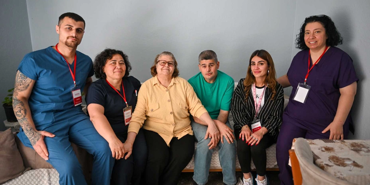 İç mekan bir odada yan yana oturan ve ayakta duran altı kişilik bir grup. En solda mavi üniformalı, dövmeli genç bir erkek; yanında lacivert üniformalı bir kadın; ortada sarı gömlekli yaşlı bir kadın; onun yanında yeşil tişörtlü bir erkek; yanında çizgili ceketli genç bir kadın ve en sağda mor üniformalı bir kadın bulunuyor. Grup üyeleri birbirlerine yakın durarak samimi bir poz veriyor, bazıları boyunlarında kurum kimlik kartları taşıyor.