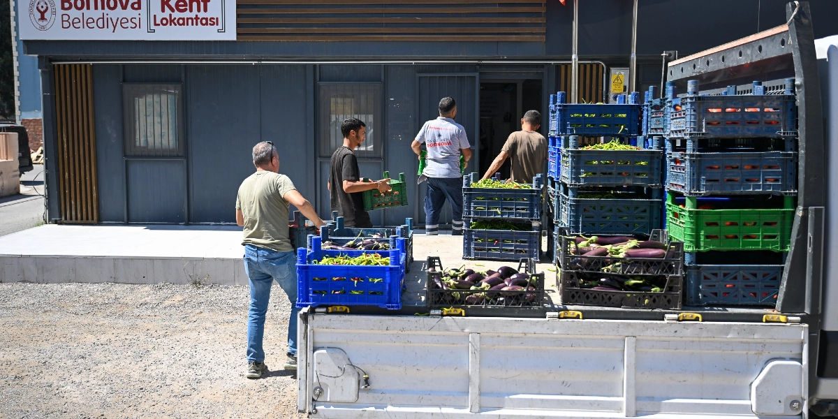 Bornova Belediyesi Kent Lokantası binasının önünde, bir kamyonetten indirilen sebze kasalarını taşıyan görevliler. Kamyonetin kasasında patlıcan ve biber dolu plastik kasalar üst üste dizilmiş halde bulunuyor, görevliler ise bu kasaları lokantaya taşıyor.