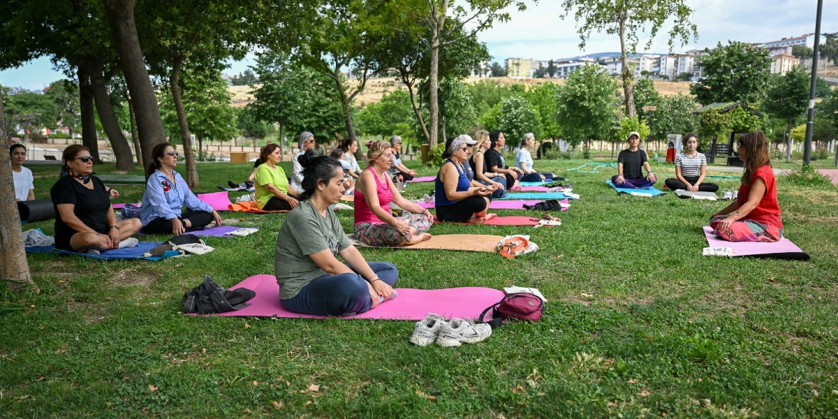 Ağaçlık bir park alanında, çimlerin üzerine serilmiş matlar üzerinde bağdaş kurarak oturan ve yoga yapan bir grup kadın. Kadınlar rahat spor kıyafetleri giymiş, eğitmen olduğu düşünülen kırmızı tişörtlü bir kadın ise grubun karşısında oturarak onlara rehberlik ediyor. Arka planda Bornova'nın yerleşim yerleri ve tepeler görünüyor.