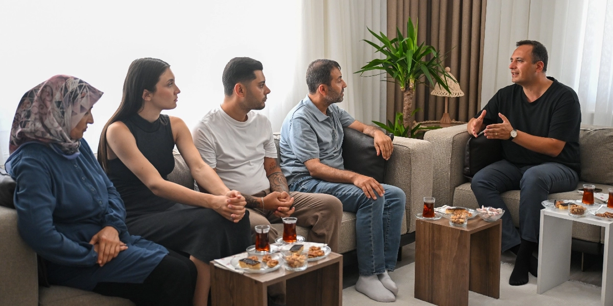 Aydınlık bir oturma odasında, bej rengi koltuklarda yan yana oturan dört kişilik bir aile ve karşısında onlara hitap eden bir erkek bulunuyor. Başörtülü bir kadın, genç bir kadın, genç bir erkek ve orta yaşlı bir adam dikkatle karşılarındaki kişiyi dinliyor. Siyah tişörtlü ve saatli adam, elleriyle jest yaparak konuşuyor. Önlerindeki ahşap sehpalarda çay bardakları ve ikramlıklar yer alıyor.