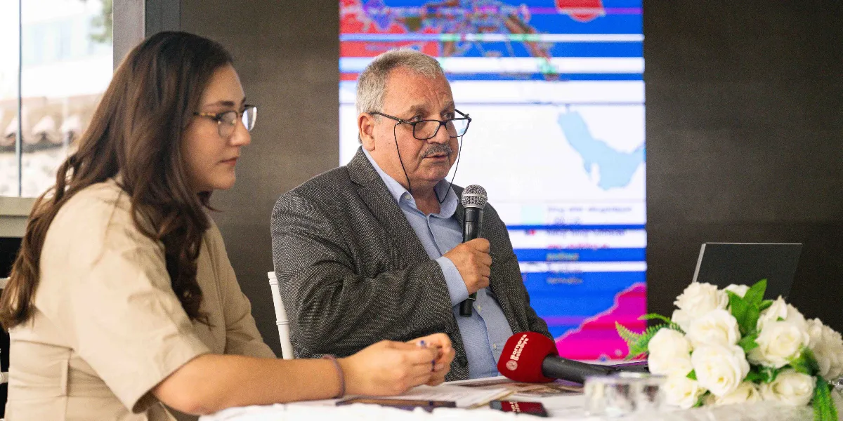 Bir toplantı masasında oturan, gözlüklü, gri ceketli ve açık mavi gömlekli orta yaşlı bir erkek, elinde mikrofon tutarak konuşma yapıyor. Yanında, bej rengi bir kıyafet giymiş, gözlüklü genç bir kadın onu dinliyor. Masanın üzerinde Bornova Belediyesi logolu bir mikrofon ve beyaz güllerden oluşan bir aranjman bulunuyor. Arka planda ise üzerinde harita görselleri bulunan büyük bir dijital ekran yer alıyor.