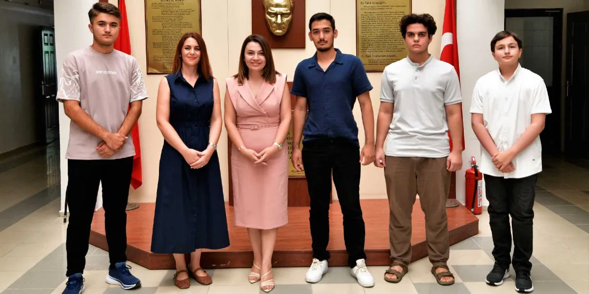 İç mekan bir salonda, arka planda Atatürk büstü ve Türk bayraklarının önünde yan yana duran iki kadın ve dört genç erkekten oluşan bir grup. Kişiler resmi bir ortamda, gülümseyerek kameraya bakıyorlar. Kadınlardan biri lacivert kolsuz bir elbise, diğeri ise pembe yakalı bir elbise giyiyor; gençler ise günlük kıyafetlerle poz veriyor.