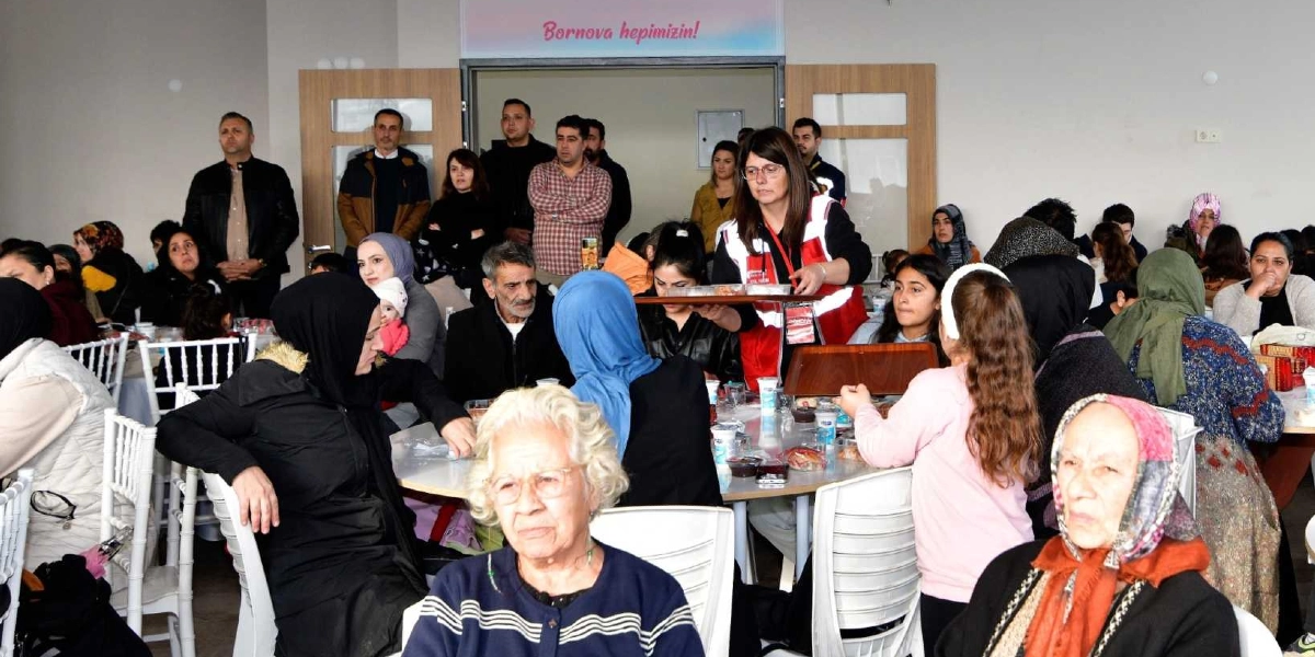Kapalı bir mekanda düzenlenen toplu yemek etkinliğinde, masalarda oturan Bornovalı vatandaşlar ve onlara yemek servisi yapan görevliler yer alıyor. Ön planda beyaz saçlı, gözlüklü yaşlı bir kadın ve başında desenli bir eşarp bulunan başka bir kadın oturuyor. Arka planda kırmızı yelekli bir görevli elindeki tepsiyle servis yaparken, duvarın üzerinde 