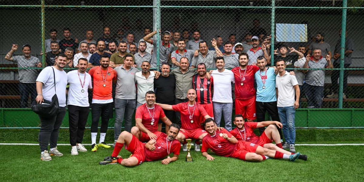 Halı sahada bir araya gelmiş, boyunlarında madalyalar olan ve önlerinde bir kupa bulunan futbol takımı oyuncuları ile onları izleyen kalabalık bir grup. Kırmızı formalar giymiş oyuncuların bir kısmı ayakta, bir kısmı ise çimlerin üzerinde oturarak poz veriyor. Arka planda tel örgüler önünde duran diğer katılımcılarla birlikte herkes neşeli bir şekilde kameraya gülümsüyor.