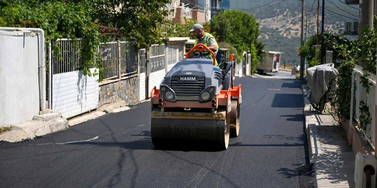 Belediye başkanı, üzerinde sarı ikaz yeleği ve şapka ile bir mahalle sokağında asfalt silindiri kullanarak yol çalışması yapıyor.