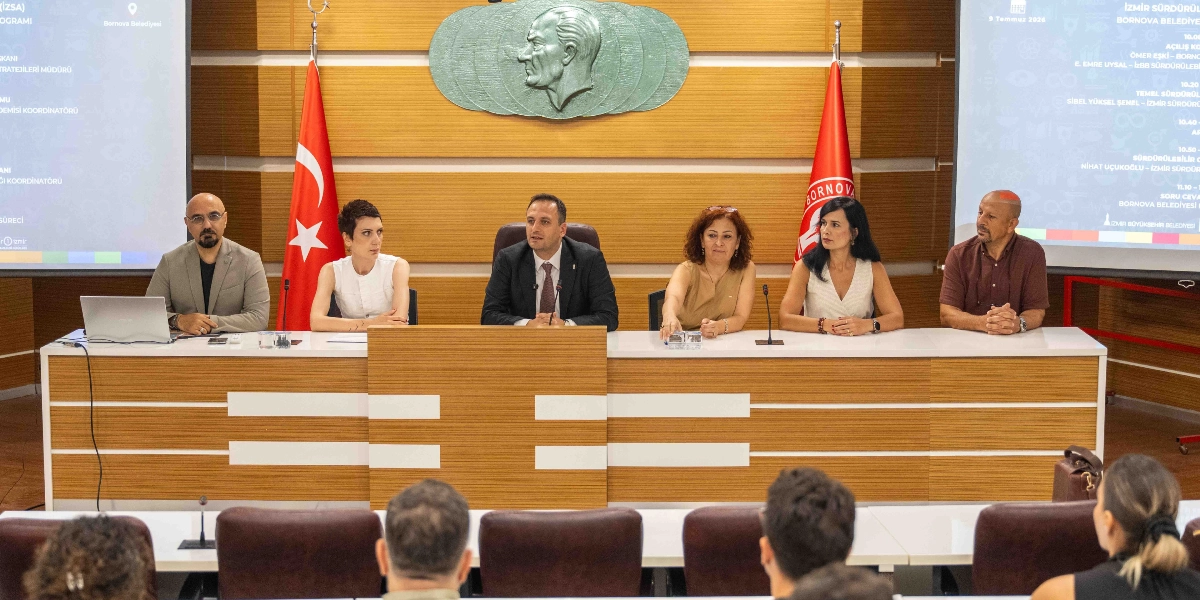 Bornova Belediyesi'nde düzenlenen bir toplantıda, uzun bir masa etrafında yan yana oturan altı kişilik bir panelist grubu konuşma yapıyor. Arka planda duvarda Atatürk silüeti, iki yanında Türk bayrağı ve Bornova Belediyesi flaması yer alıyor. Masanın iki ucundaki projeksiyon ekranlarında 