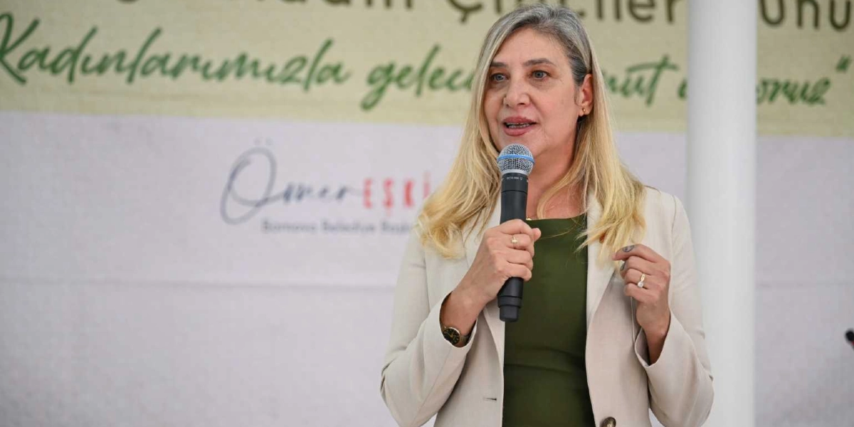 Sarı saçlı, açık renk ceket ve haki yeşil bir bluz giyen bir kadın, elinde mikrofonla konuşma yapıyor. Arka planda üzerinde 