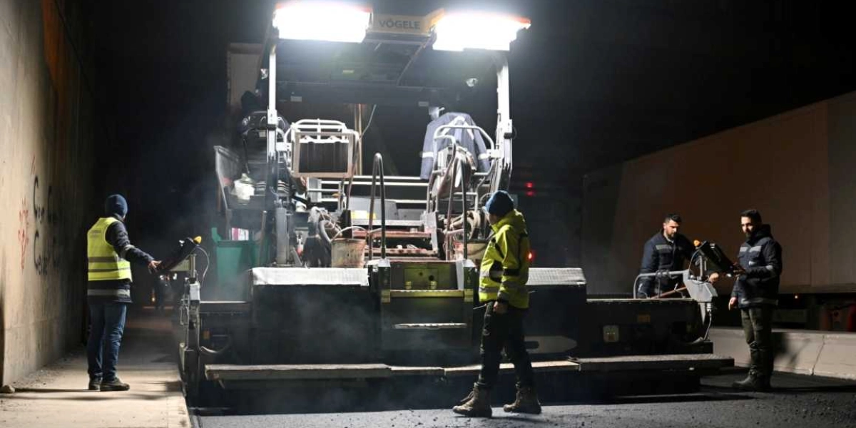 Gece saatlerinde bir tünel veya alt geçit içerisinde çalışan işçiler ve asfalt serme makinesi. Sarı reflektörlü yelek giymiş dört işçi, güçlü aydınlatmalarla donatılmış Vögele marka asfalt serim makinesinin çevresinde çalışıyor. Makineden yükselen dumanlar ve yeni serilmiş asfaltın dokusu görülüyor.