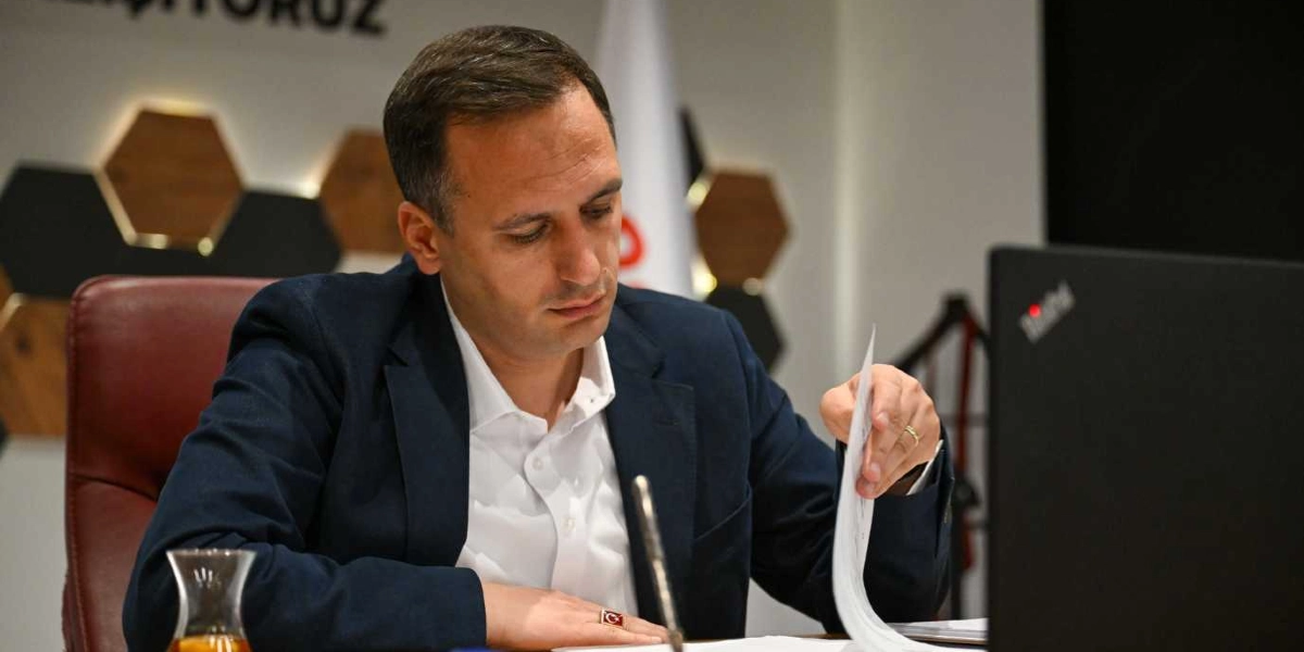Lacivert ceket ve beyaz gömlek giymiş, parmağında Türk bayraklı bir yüzük bulunan bir erkek, ofis ortamında masasında oturarak elindeki belgeleri inceliyor. Önünde bir dizüstü bilgisayar ve ince belli bir bardakta çay bulunuyor. Arka planda ise altıgen dekoratif paneller yer alıyor.