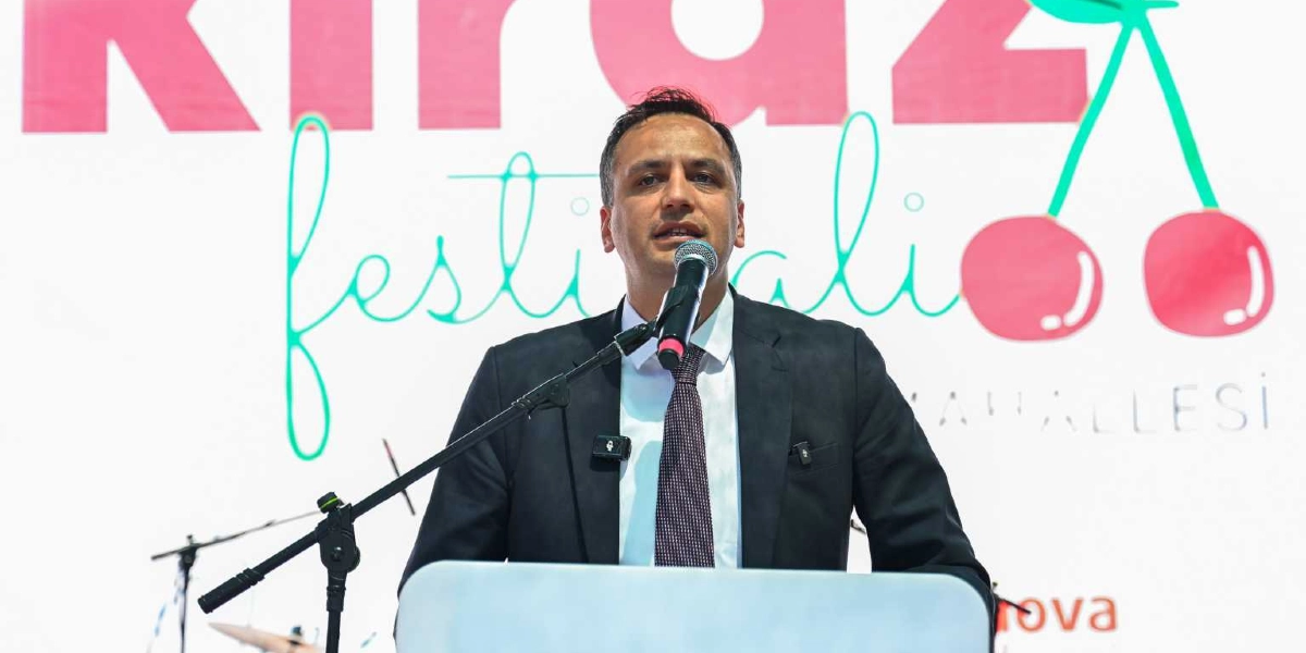 Koyu renk takım elbiseli ve kravatlı bir erkek, önündeki kürsüde bulunan mikrofona konuşma yapıyor. Arka planda üzerinde 