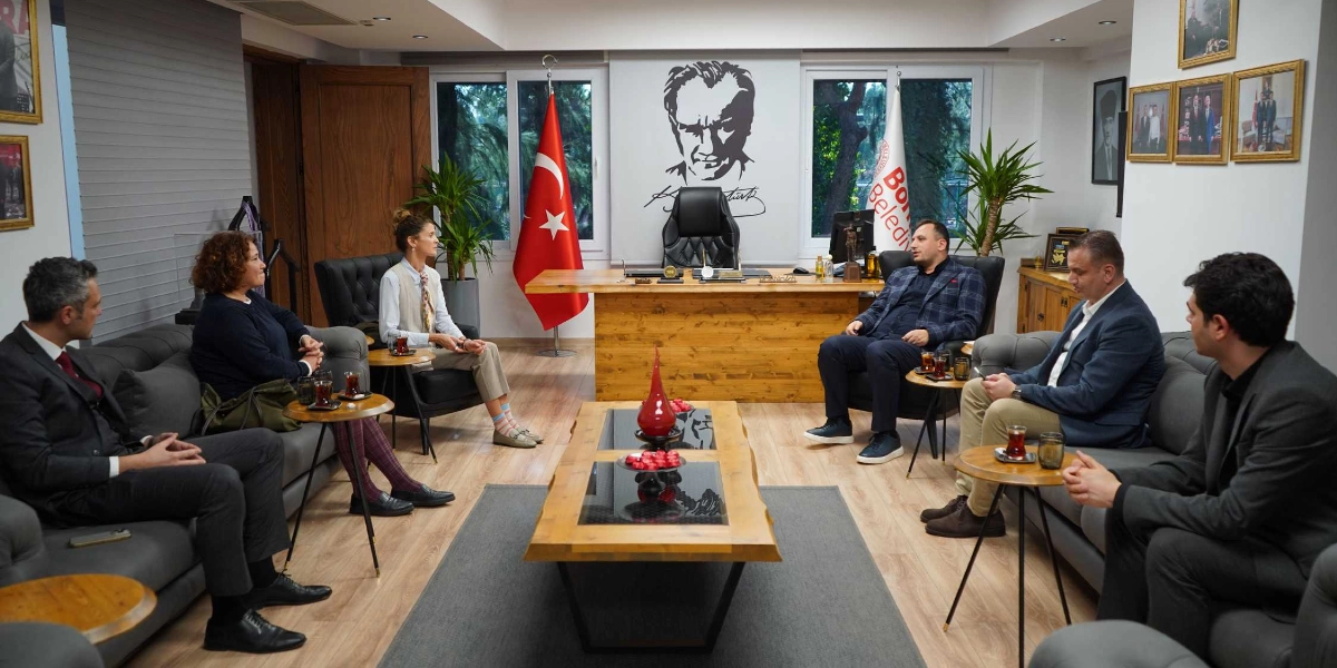 Bornova Belediyesi makam odasında, Bornova Belediye Başkanı ve İnci Vakfı temsilcilerinin yer aldığı bir toplantı. Odada, ortadaki ahşap bir sehpanın etrafındaki gri koltuklarda oturan altı kişilik bir grup bulunuyor. Arka planda duvarda Atatürk portresi, yanında Türk bayrağı ve Bornova Belediyesi bayrağı yer alıyor. Katılımcılar karşılıklı oturmuş, sohbet ediyor ve önlerindeki küçük sehpalarda çay bardakları bulunuyor.
