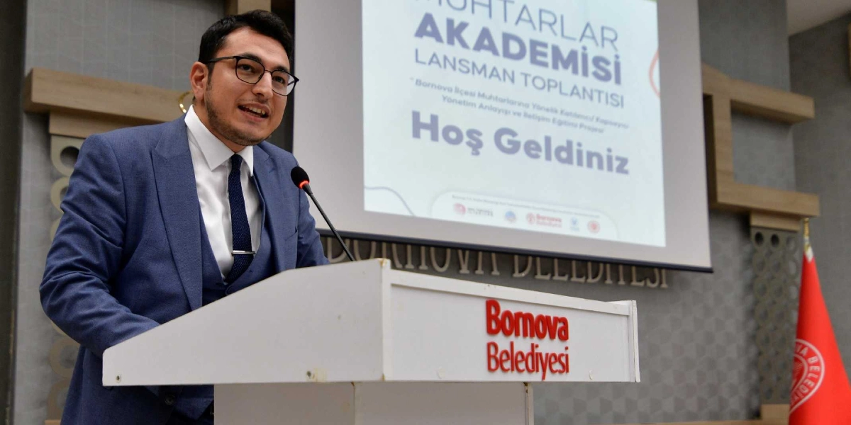 Mavi takım elbiseli, gözlüklü bir konuşmacı, üzerinde Bornova Belediyesi logosu bulunan bir kürsüde mikrofon başında hitap ediyor. Arkasındaki projeksiyon perdesinde Muhtarlar Akademisi Lansman Toplantısı Hoş Geldiniz yazısı ve Bornova Belediyesi'nin kurumsal logoları yer alıyor.