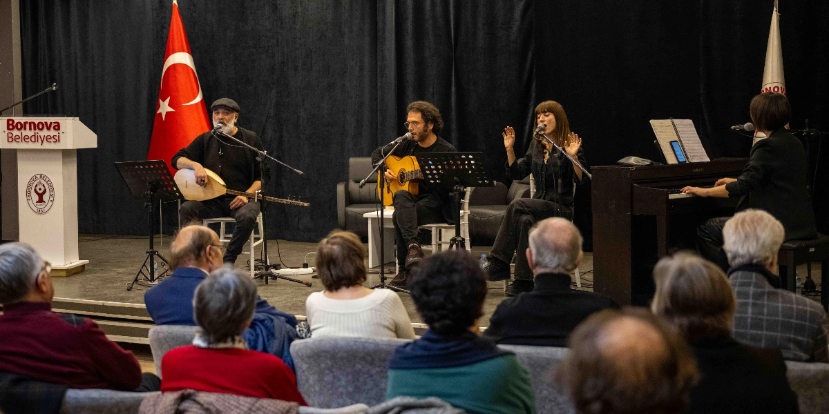Bornova Belediyesi'ne ait bir sahnede, siyah perdeler önünde dört kişilik bir müzik grubu performans sergiliyor. Solda bağlama çalan şapkalı bir erkek, yanında gitar çalan bir erkek, onun yanında şarkı söyleyen bir kadın ve en sağda piyano çalan bir kadın yer alıyor. Sahnenin sol tarafında üzerinde Bornova Belediyesi logosu bulunan bir kürsü ve Türk bayrağı bulunuyor. İzleyiciler, sahnedeki müzisyenleri karşıdan izliyor.