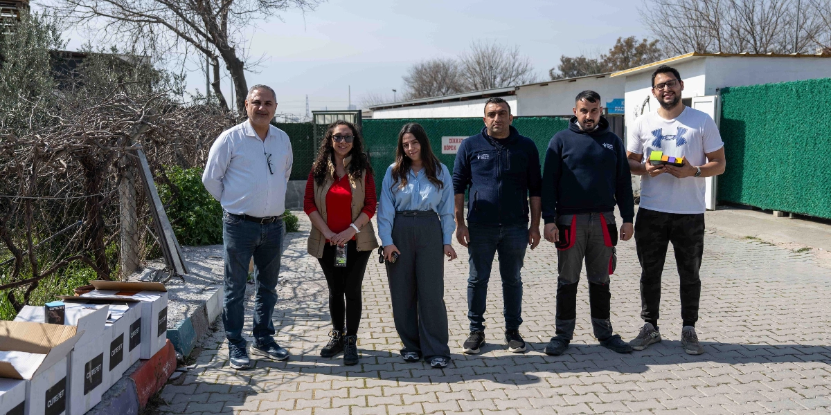 Açık havada, parke taşlı bir alanda yan yana duran altı kişilik bir grup. Grubun sol tarafında, yerde üzerinde mama markası yazılı olan birkaç kutu bulunuyor. Sağ taraftaki kişi elinde renkli küçük kutulardan oluşan bir set tutuyor. Arka planda yeşil çitlerle çevrili bir yapı ve ağaçlar yer alıyor.