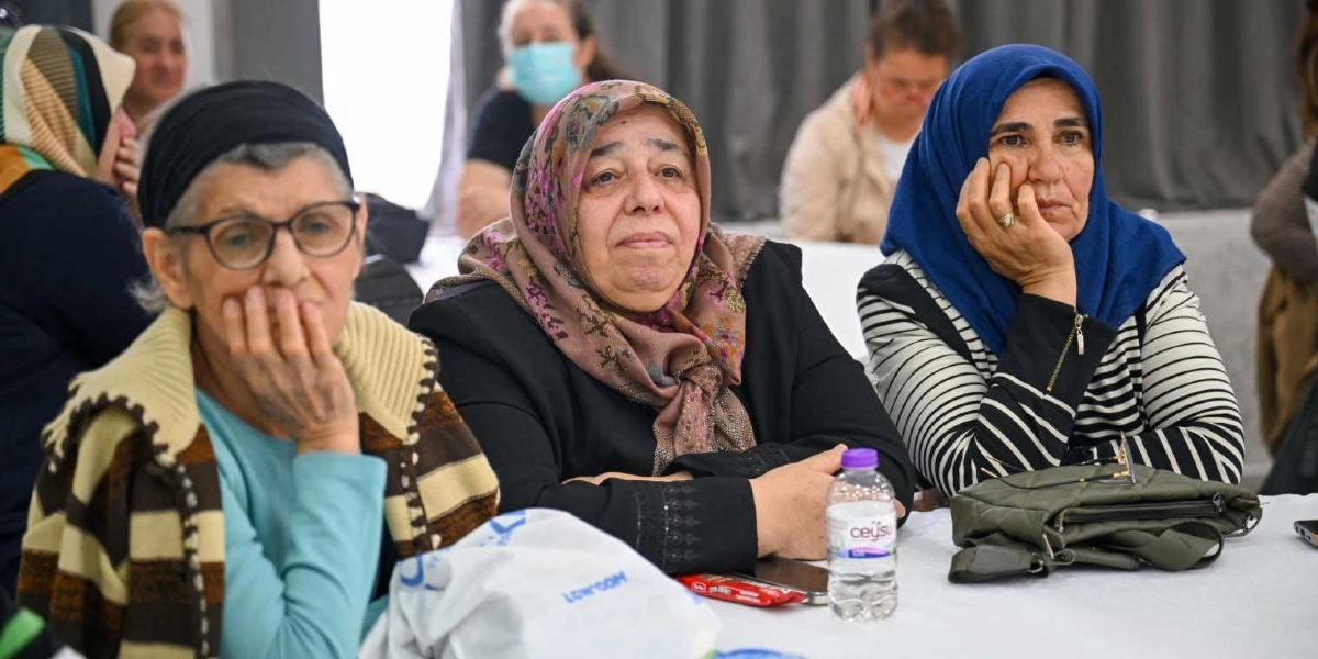 Bir etkinlikte yan yana oturmuş, dikkatle dinleyen başörtülü kadınlar. Önlerinde beyaz bir masa örtüsü üzerinde su şişesi ve kişisel eşyalar bulunuyor. Kadınlar farklı renklerde başörtüleri ve günlük kıyafetleriyle, odaklanmış bir şekilde karşıya bakıyorlar.
