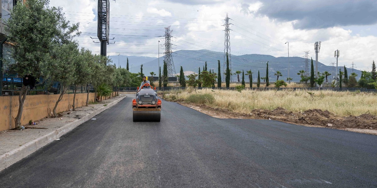 Bir operatörün kullandığı turuncu renkli silindir, yeni serilmiş pürüzsüz asfalt üzerinde ilerliyor. Yolun sol tarafında ağaçlar ve bir duvar, sağ tarafında ise boş bir arazi ve arka planda dağ manzarası ile elektrik direkleri bulunuyor.