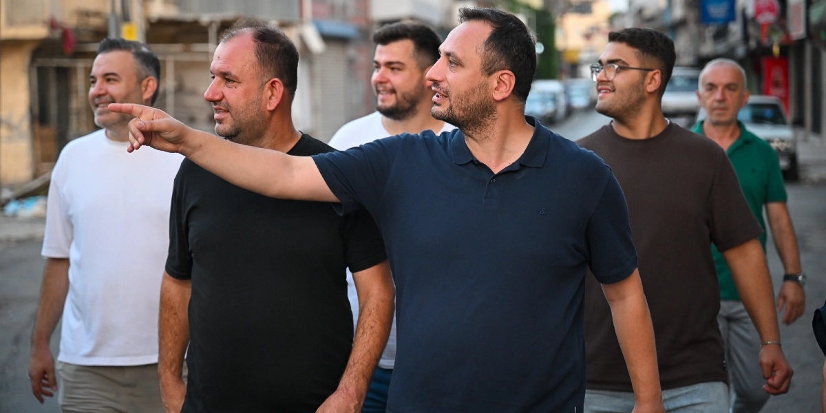 Çamdibi sokaklarında yürüyen bir grup erkek, lacivert polo yaka tişörtlü bir adamın eliyle işaret ettiği yöne doğru bakıyor. Gruptaki diğer kişiler beyaz, siyah, kahverengi ve yeşil tişörtler giymiş durumda; arka planda ise yerleşim yeri ve araçlar görülüyor.