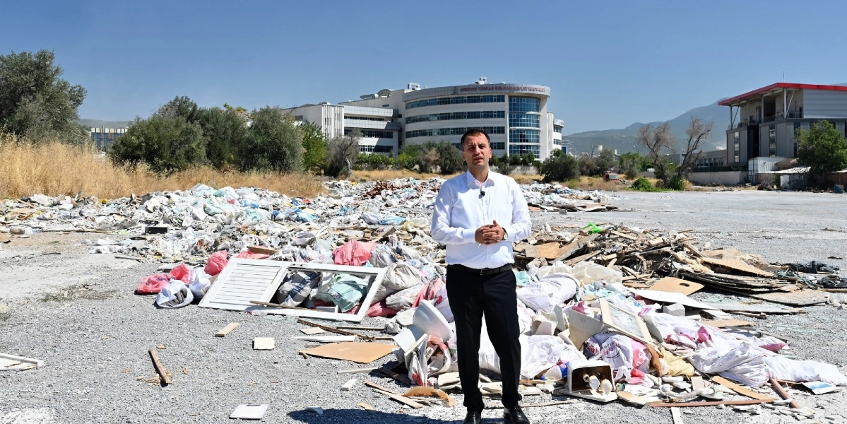 Beyaz gömlekli ve siyah pantolonlu bir erkek, etrafı moloz yığınları ve inşaat atıklarıyla dolu boş bir arazide durarak konuşma yapıyor. Arka planda Bornova Türkan Özilhan Devlet Hastanesi binası ve açık bir gökyüzü görülüyor.