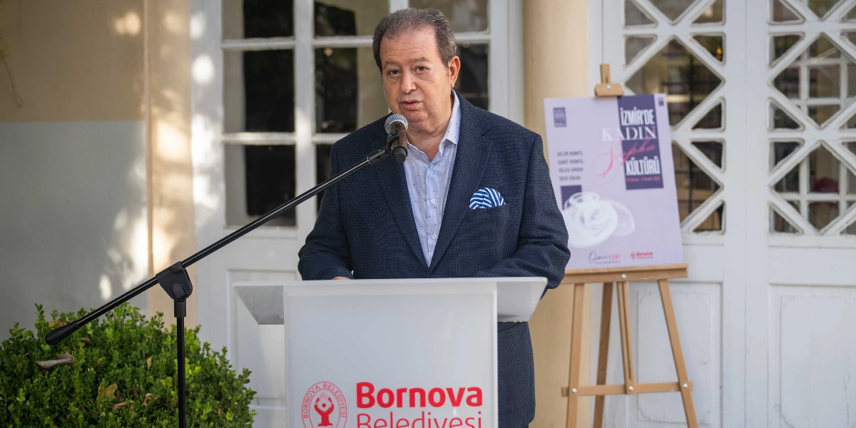 Koyu renk ceket ve açık renk gömlek giymiş orta yaşlı bir erkek, üzerinde Bornova Belediyesi logosu bulunan bir kürsüde mikrofon başında konuşma yapıyor. Arka planda, üzerinde 