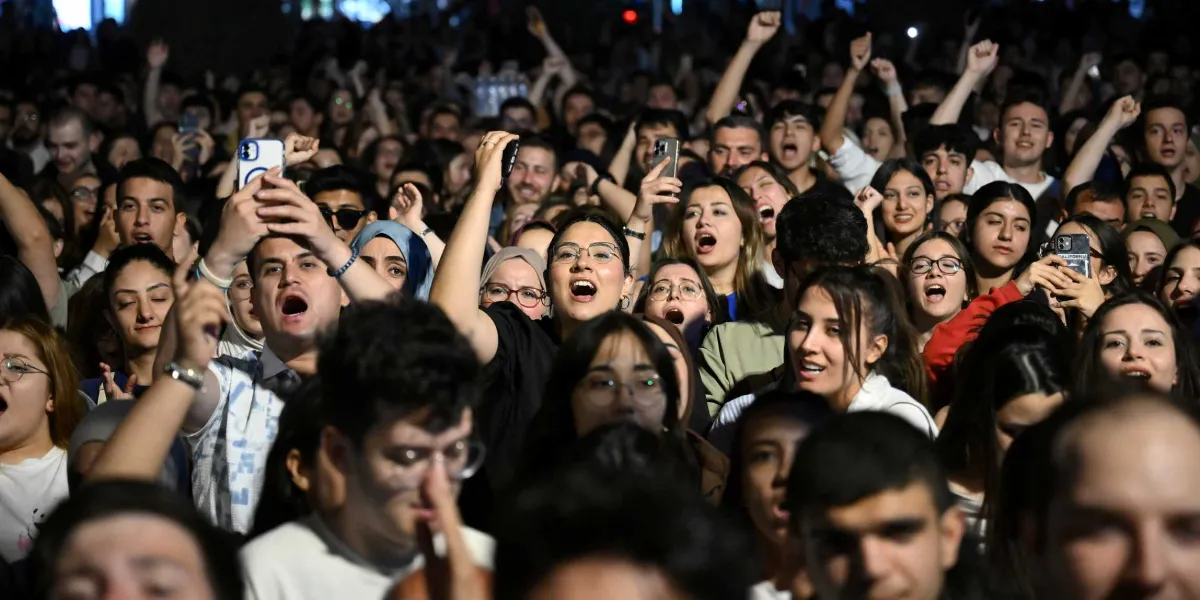 Gece vakti açık havada düzenlenen bir etkinlikte, coşkuyla şarkı söyleyen veya slogan atan, ellerini havaya kaldırmış ve bazıları cep telefonlarıyla kayıt yapan kalabalık bir genç grubu.