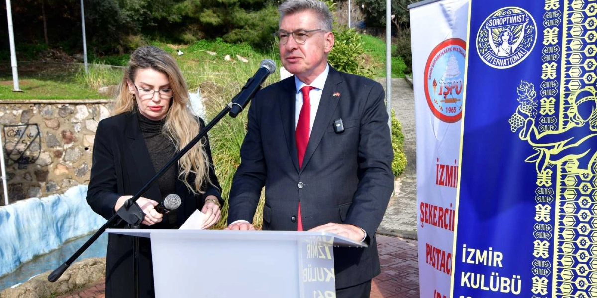 Koyu renk ceket giymiş, gözlüklü bir erkek kürsüde konuşma yaparken, yanında siyah kıyafetli sarı saçlı bir kadın mikrofon tutuyor. Arka planda üzerinde İzmir Şekerciler ve Pastacılar Odası ile Soroptimist International logolarının bulunduğu dikey bir pankart yer alıyor.