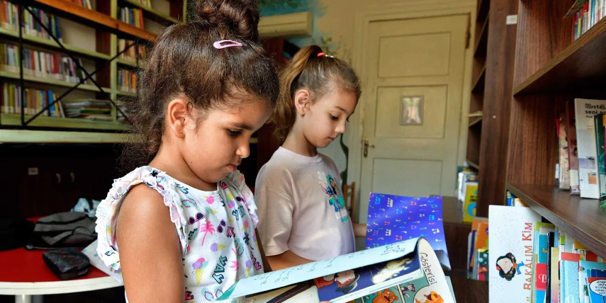 Kütüphane ortamında kitap raflarının önünde duran iki kız çocuğu, ellerindeki kitapları dikkatle inceliyor. Ön plandaki çocuk beyaz desenli bir tişört giymiş, saçlarını tepeden toplamış ve pembe bir toka takmış; yanındaki arkadaşı ise üzerinde çizgi film karakteri baskısı olan açık renkli bir tişört giyiyor. Arkalarında kitaplarla dolu raflar ve kapalı bir kapı bulunuyor.