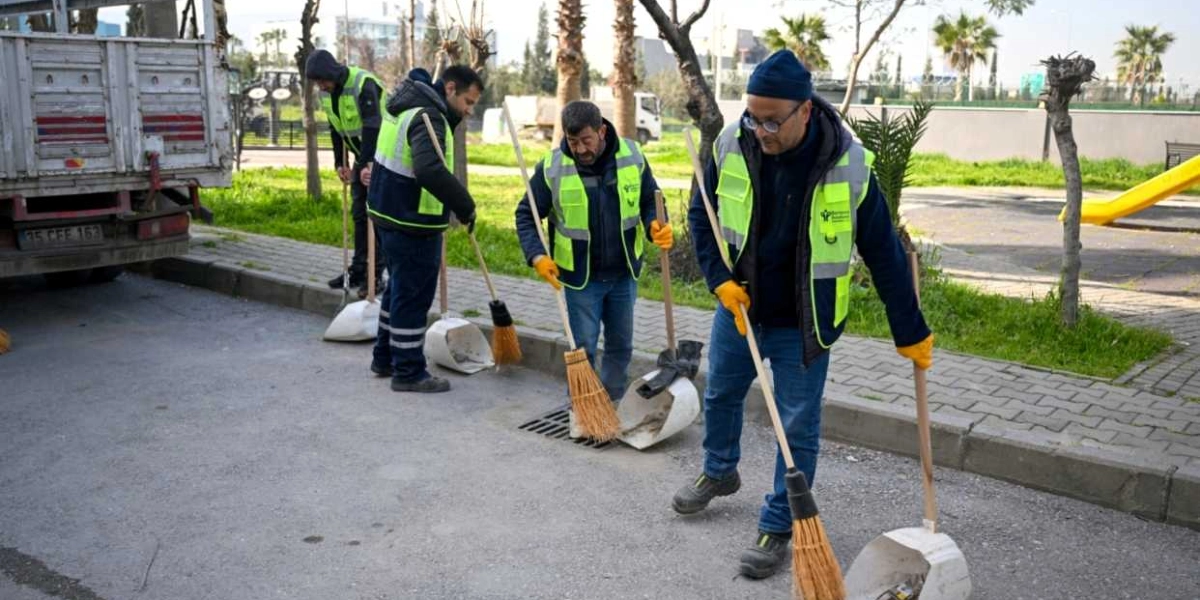 Üzerlerinde Bornova Belediyesi logolu fosforlu yelekler bulunan dört belediye çalışanı, bir parkın kenarındaki asfalt yolda ellerinde süpürge ve faraşlarla temizlik yapıyor. Çalışanlardan biri ön planda, diğerleri ise arkasında hizalanmış şekilde yer alıyor; sol tarafta ise temizlik çalışmalarına destek veren bir kamyonun arka kısmı görünüyor.