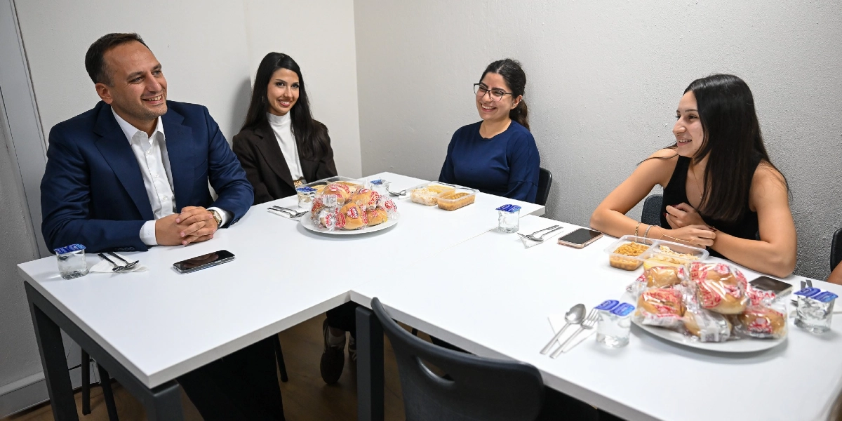 Lacivert ceketli bir erkek, beyaz bir masa etrafında oturan üç genç kadınla sohbet ediyor. Masanın üzerinde tabaklarda paketli ekmekler, plastik kaplarda yemekler ve küçük su bardakları bulunuyor. Herkesin yüzünde gülümseme olan samimi bir ortamda, öğrenciler ve belediye başkanı yemek eşliğinde bir araya gelmiş durumda.