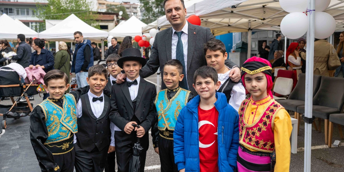 Bornova Belediye Başkanı, 23 Nisan Ulusal Egemenlik ve Çocuk Bayramı etkinlikleri kapsamında geleneksel kıyafetler ve şık takım elbiseler giymiş bir grup çocukla birlikte açık havada poz veriyor. Çocuklardan bazıları yöresel halk oyunu kostümleri, bazıları ise papyonlu takım elbiseler giymiş durumda; bir çocuk ise üzerinde Türk bayrağı baskılı kırmızı bir tişört ve mavi bir montla yer alıyor. Arka planda beyaz çadırlar ve etkinliğe katılan diğer insanlar görülüyor.