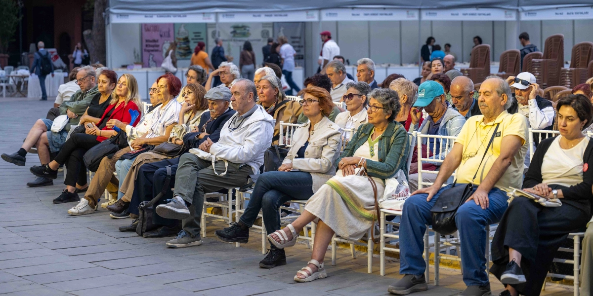 Açık havada düzenlenen bir etkinlikte, beyaz sandalyelerde yan yana oturarak dikkatle dinleyen, çoğunluğu orta ve ileri yaş grubundan oluşan izleyici kitlesi. Arka planda ise beyaz çadırlar altında kurulmuş stantlar ve kitap fuarı görünümü yer alıyor.