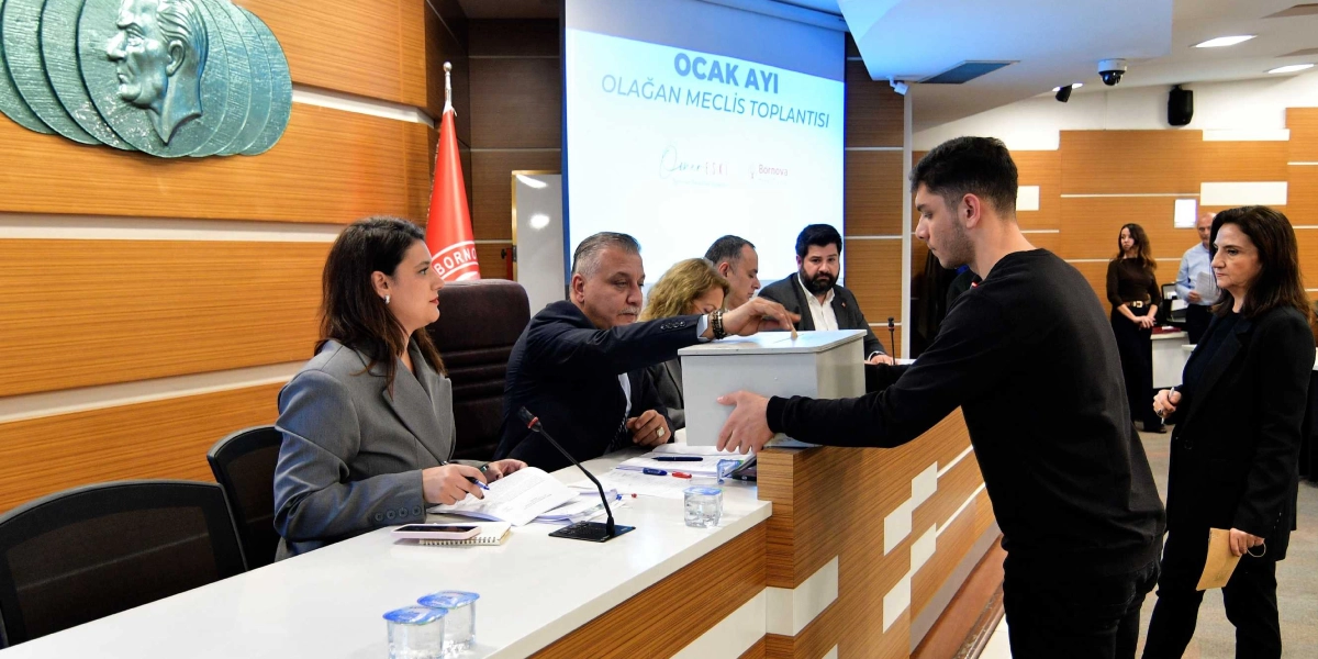 Bornova Belediyesi meclis salonunda düzenlenen Ocak Ayı Olağan Meclis Toplantısı'nda, bir genç elindeki zarfı sandığa atarken, masada oturan yetkililer süreci takip ediyor. Arka planda 