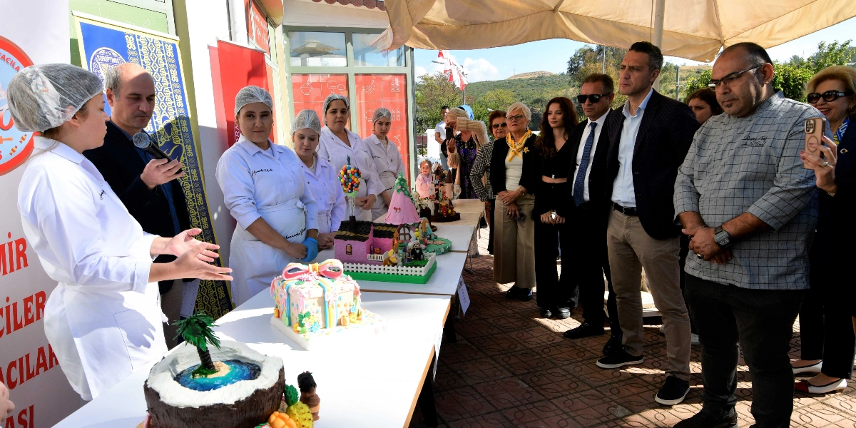 Açık havada düzenlenen bir etkinlikte, beyaz önlük ve bone giymiş pastacılık kursiyerleri, hazırladıkları süslü pasta tasarımlarını sergiliyor. Ön planda palmiye ağacı figürlü bir pasta, hediye paketi şeklinde bir pasta ve üzerinde balonlar bulunan ev figürlü bir pasta yer alıyor. Kursiyerlerden biri elinde mikrofonla konuşurken, diğerleri ve katılımcılar masanın etrafında toplanmış, çalışmaları inceliyor. Arka planda Soroptimist Kulübü'ne ait olduğu anlaşılan logolu bir afiş ve etkinlik alanını gölgeleyen büyük bir şemsiye bulunuyor.