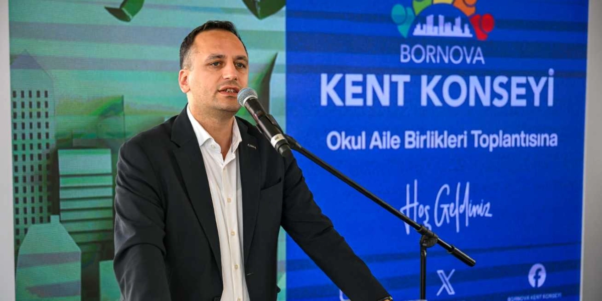 Koyu renk ceket ve beyaz gömlek giymiş bir erkek, önündeki mikrofona konuşma yapıyor. Arkasında üzerinde 