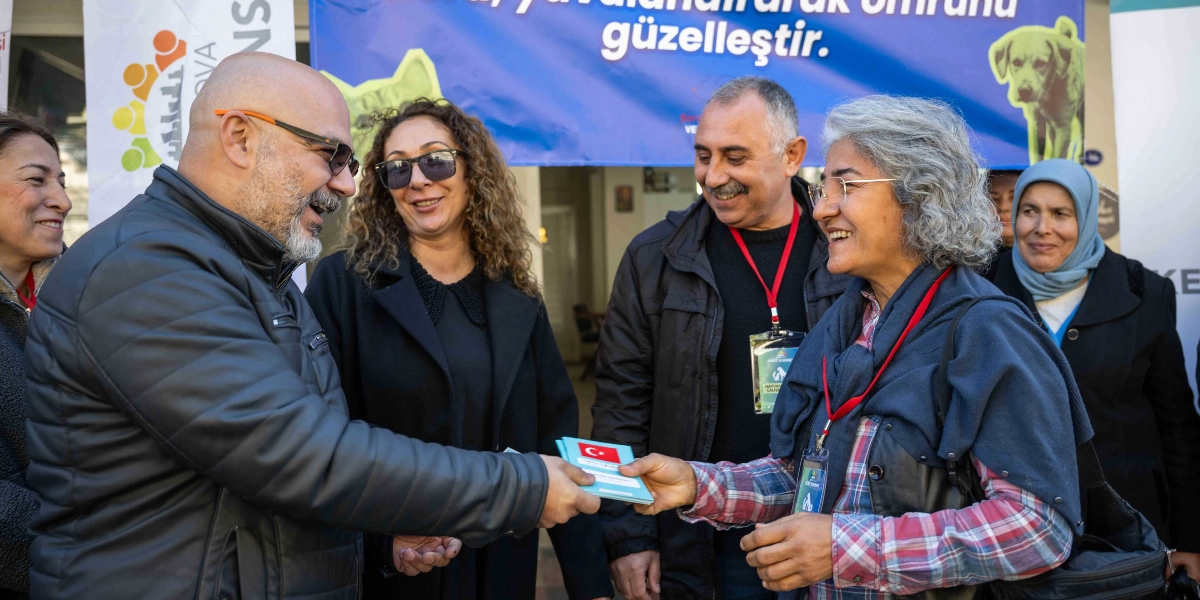 Bornova Kent Konseyi tarafından düzenlenen hayvan sahiplendirme etkinliğinde, sakallı ve gözlüklü bir erkek, üzerinde Türk bayrağı bulunan bir sahiplendirme karnesini gri saçlı bir kadına teslim ediyor. Etkinliğe katılan diğer kişiler gülümseyerek bu anı izliyor. Arka planda üzerinde 