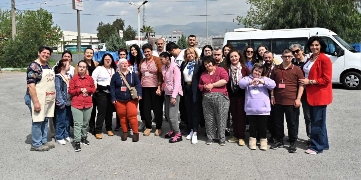 Açık havada, asfalt bir alanda bir araya gelmiş, aralarında özel gereksinimli bireylerin ve eğitmenlerin bulunduğu kalabalık bir grup. Grubun en solunda üzerinde 