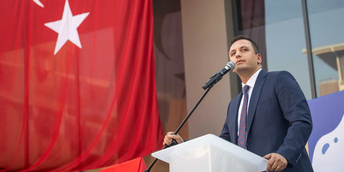 Koyu renk takım elbiseli ve çizgili kravatlı bir erkek, kürsüde mikrofon başında konuşma yapıyor. Arka planda büyük bir Türk bayrağı asılı duruyor.