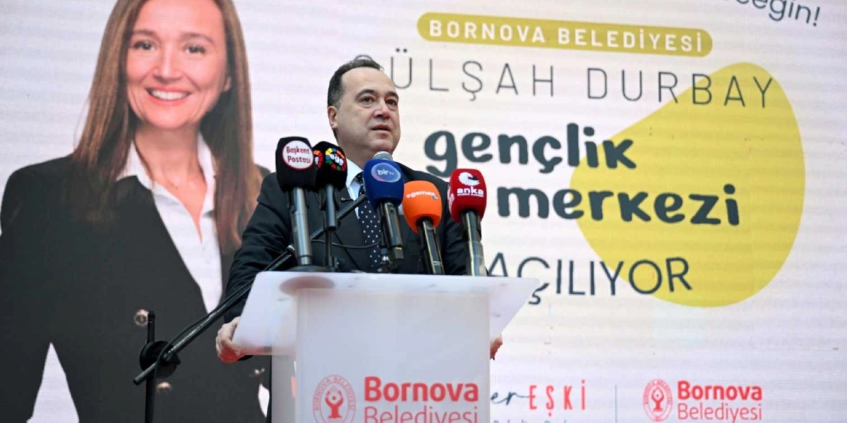 Bornova Belediye Başkanı Ömer Eşki, kürsüde konuşma yaparken görülüyor. Kürsünün önünde çeşitli basın kuruluşlarına ait mikrofonlar yer alıyor. Arka planda ise üzerinde 