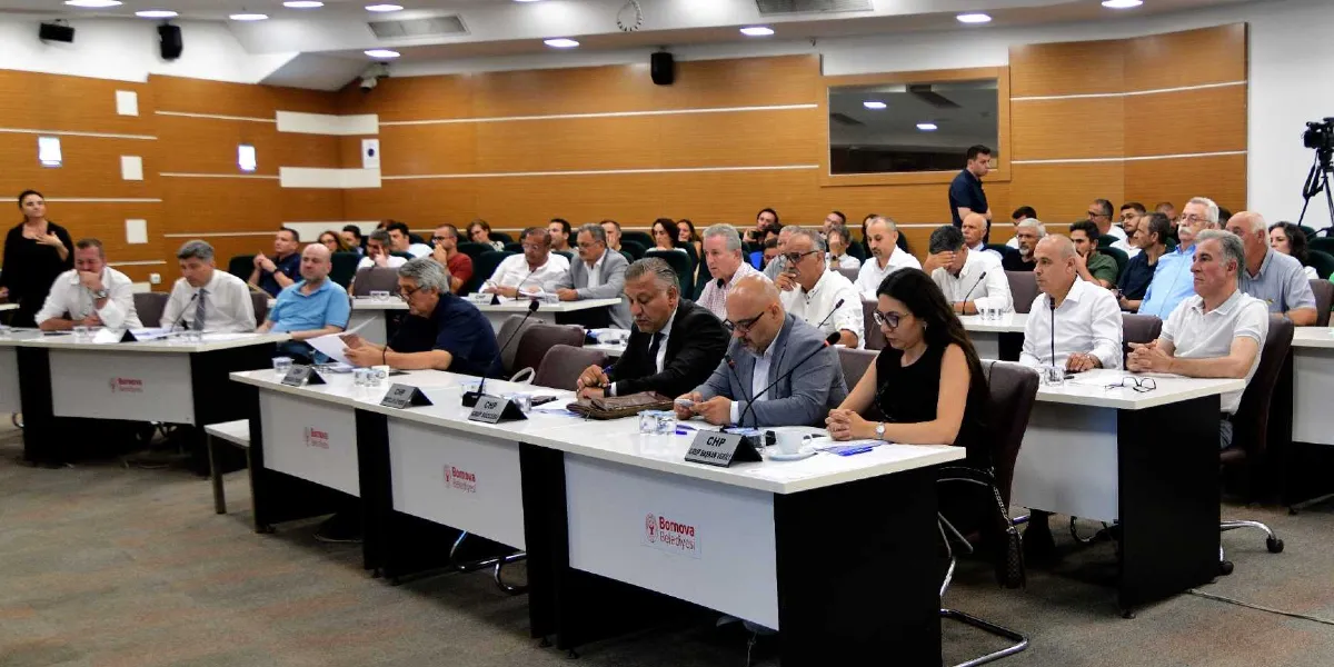 Bornova Belediyesi meclis salonunda düzenlenen bir toplantıda, masalarda oturan çok sayıda katılımcı yer alıyor. Katılımcılar önlerindeki mikrofonlar ve isimliklerle toplantıyı takip ederken, masaların üzerinde Bornova Belediyesi logoları bulunuyor. Salonun ahşap panellerle kaplı duvarları ve düzenli oturma düzeniyle resmi bir toplantı ortamı görülüyor.