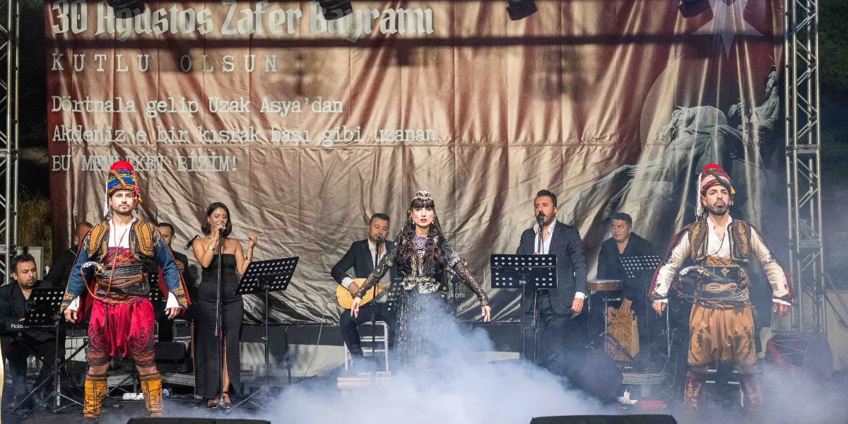 30 Ağustos Zafer Bayramı kutlamaları kapsamında sahnede geleneksel kıyafetli iki erkek dansçı, yöresel kıyafetli bir kadın solist ve müzisyenlerden oluşan bir grup performans sergiliyor. Arka planda üzerinde 