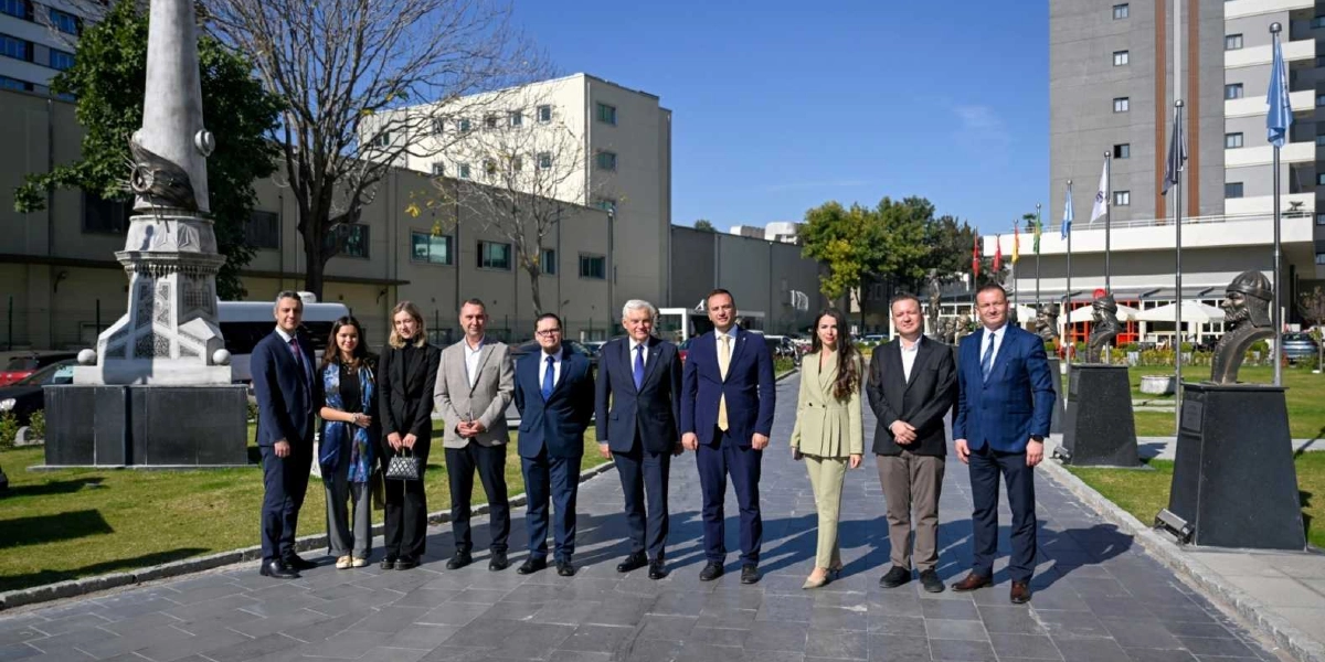 Açık havada, taş döşeli bir yolda yan yana duran on kişilik resmi heyet. Grubun solunda yüksek bir anıt, sağında ise kaideler üzerinde büstler ve arka planda çeşitli bayraklar yer alıyor. Çoğunluğu takım elbiseli olan grup, Bornova ve Białystok arasındaki dostluk ilişkilerini geliştirmek amacıyla bir araya gelmiş yetkililerden oluşuyor. Arka planda modern binalar ve ağaçlar görülüyor.