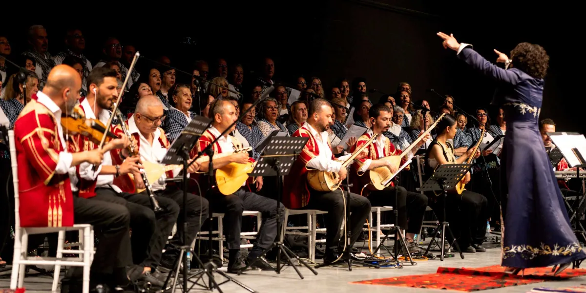 Karanlık bir sahnede, geleneksel kırmızı işlemeli ceketler giymiş müzisyenlerden oluşan bir orkestra ve arkalarında geniş bir koro yer alıyor. Müzisyenler bağlama ve keman gibi enstrümanlar çalarken, sahnenin sağ tarafında sırtı dönük, lacivert uzun bir elbise giymiş kadın şef orkestrayı yönetiyor. Arka planda ise beyaz desenli kıyafetler giymiş kalabalık bir koro grubu, ellerindeki notalarla şarkı söylüyor.