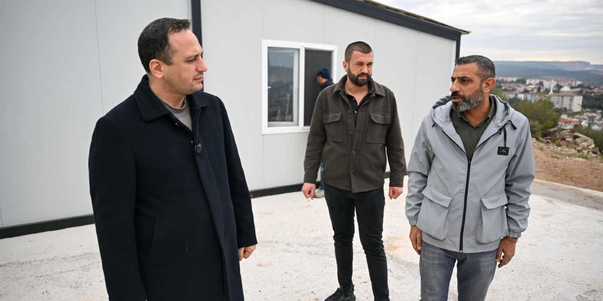 Belediye başkanı olduğu anlaşılan koyu renk paltolu bir erkek, İZYUVA yerleşkesindeki konteyner evlerin önünde iki kişiyle sohbet ediyor. Konuştuğu kişilerden biri koyu renk ceket ve pantolon giymiş, diğeri ise gri bir ceket ve kot pantolonla ayakta duruyor. Arka planda ise yerleşimin bulunduğu tepelik alan ve uzaktan görünen şehir manzarası yer alıyor.