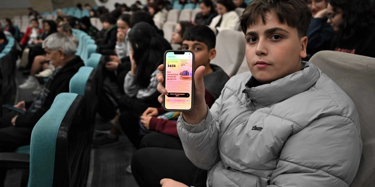 Konferans salonunda oturan gri şişme montlu bir genç, elinde tuttuğu akıllı telefonu kameraya doğru gösteriyor. Telefon ekranında 