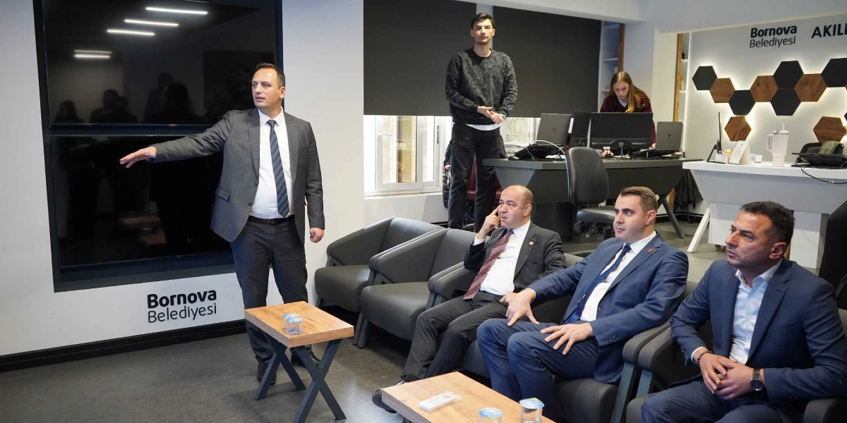 CHP Genel Başkan Yardımcısı Özgür Karabat, Bornova Belediyesi'ne ait modern bir ofis ortamında, duvardaki büyük ekranı işaret ederek bir sunum yapıyor. Karabat'ın karşısında, siyah deri koltuklarda oturan üç kişi dikkatle onu dinliyor. Arka planda, çalışma masasında bilgisayar başında çalışan bir kadın ve ayakta duran bir genç yer alıyor. Duvarlarda 