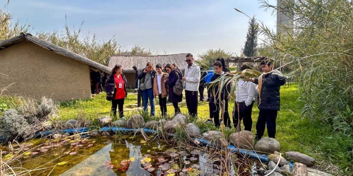 Açık havada, sazlıklarla çevrili küçük bir göletin etrafında toplanmış, aralarında öğrencilerin ve yetişkinlerin bulunduğu bir grup insan. Arka planda sazdan çatılı, kerpiç görünümlü tarihi tarzda yapılar yer alıyor. Grup, göletin kenarında durarak çevreyi inceliyor; bazı öğrenciler bitkileri gözlemliyor.