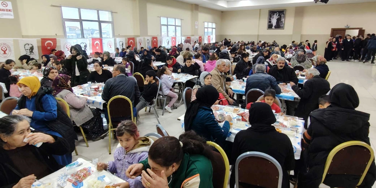 Geniş bir yemek salonunda, Bornova Belediyesi logolu bayraklar ve Türk bayraklarıyla süslenmiş masalarda oturan kalabalık bir grup insan, birlikte yemek yiyor. Salonun genelinde farklı yaş gruplarından vatandaşlar masalarda otururken, arka planda Bornova Belediye Başkanı Ömer Eşki, katılımcılarla sohbet ederek masaları dolaşıyor.