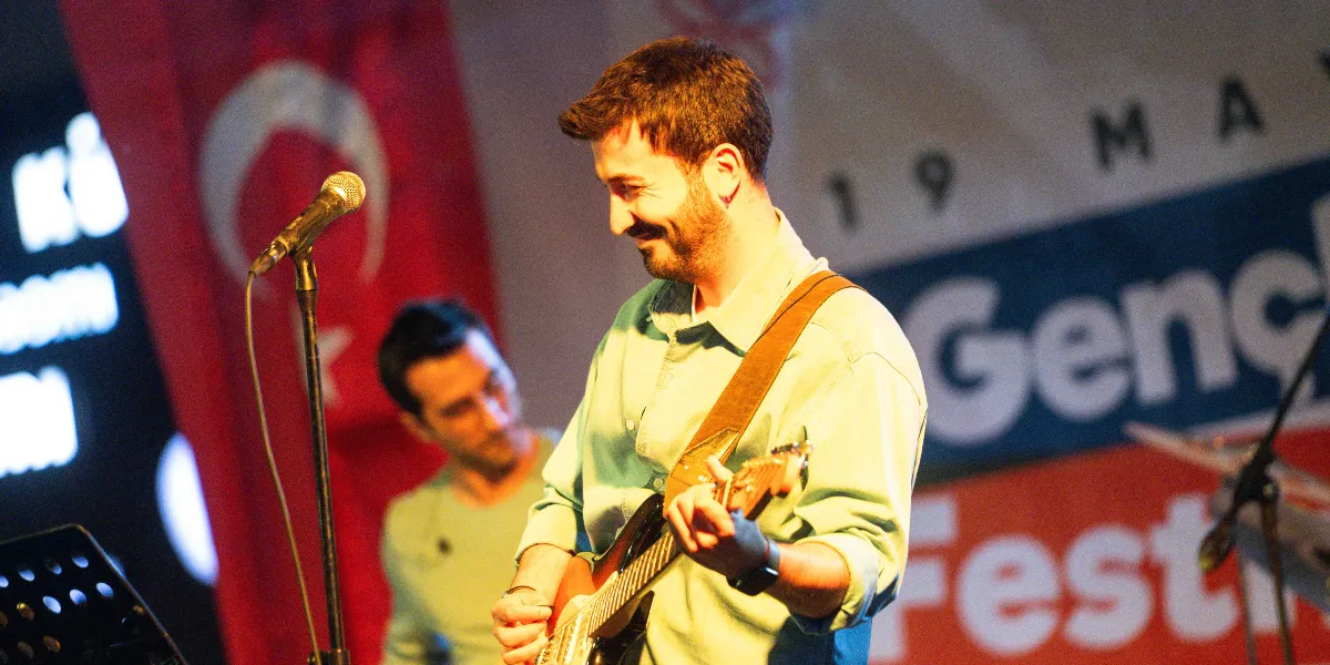 Sahne üzerinde açık renkli bir gömlek giymiş, gülümseyerek elektro gitar çalan sakallı bir müzisyen ve arkasında ona eşlik eden bir başka müzisyen yer alıyor. Arka planda Türk bayrağı ve üzerinde 