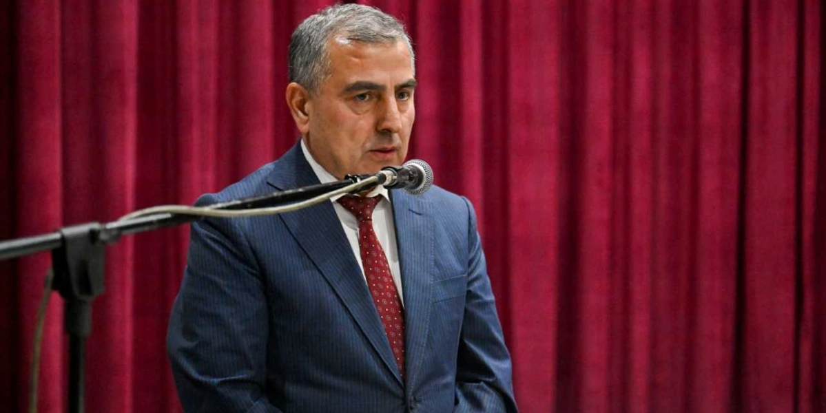 Koyu renkli takım elbise ve desenli bordo kravat giymiş, kısa saçlı orta yaşlı bir erkek, önündeki mikrofona konuşma yapıyor. Arka planda ise boydan boya kırmızı kadife bir perde bulunuyor.
