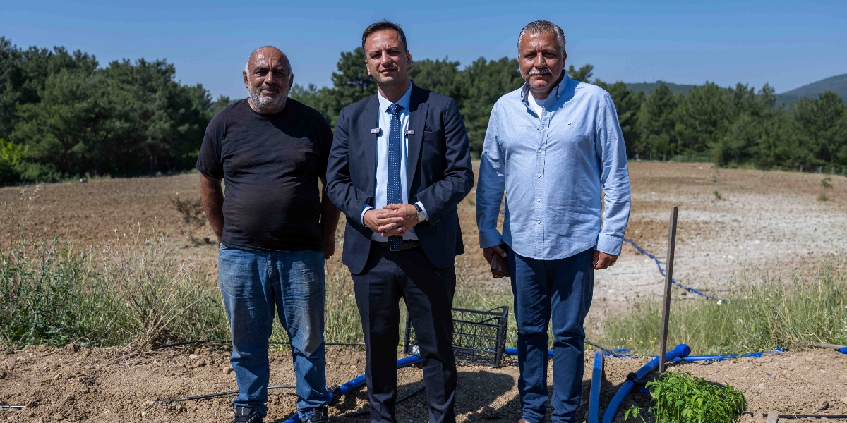 Açık havada, ağaçlık bir alanın önündeki toprak arazide yan yana duran üç erkek. Ortada lacivert takım elbiseli ve kravatlı bir kişi, solunda siyah tişörtlü ve kot pantolonlu bir kişi, sağında ise açık mavi gömlekli ve koyu renk pantolonlu bir kişi yer alıyor. Ön planda toprağa serilmiş mavi sulama boruları ve yeni dikilmiş küçük yeşil fideler görülüyor.